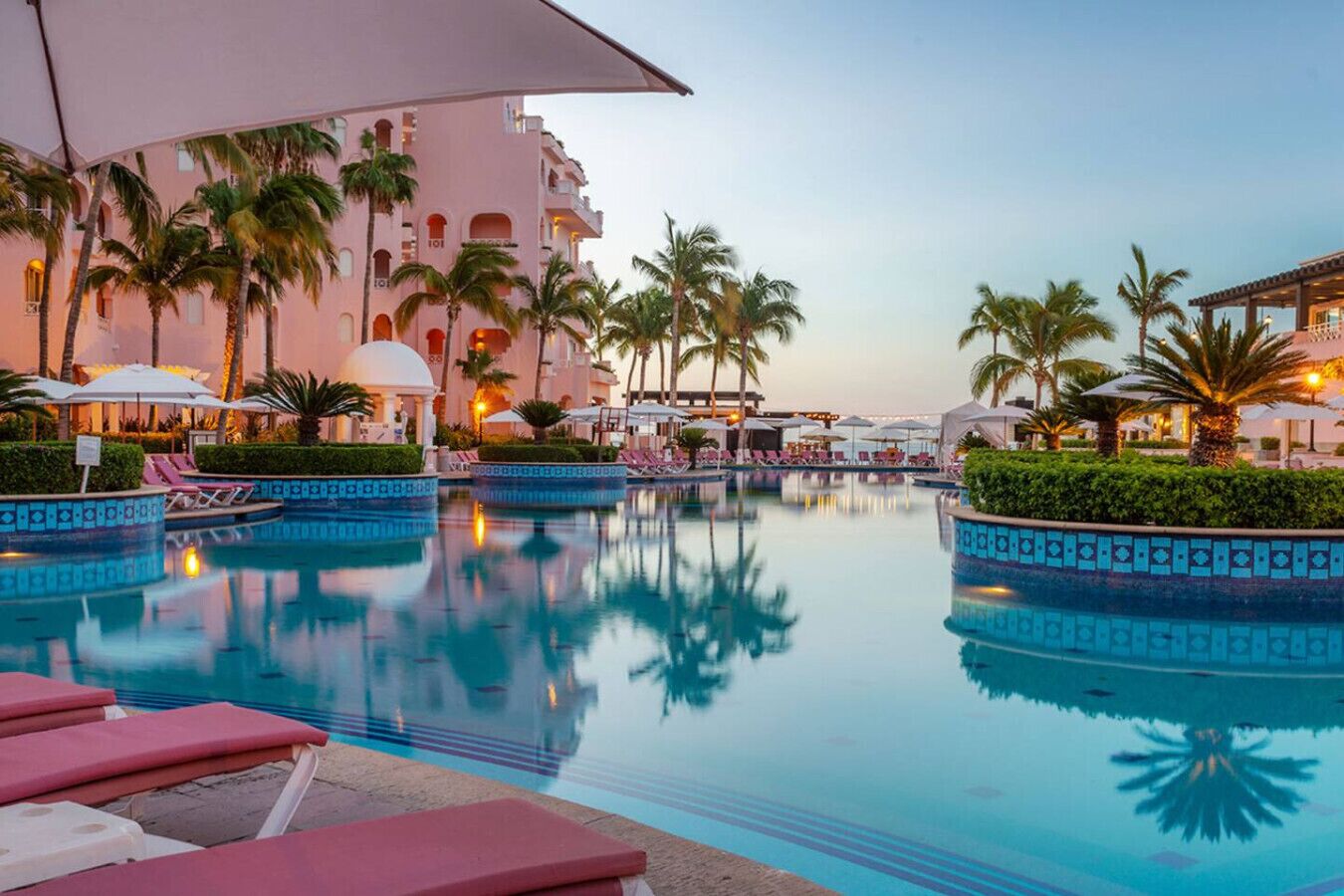 A hotel pool area with lounge chairs, palm trees, and a large building in the background.