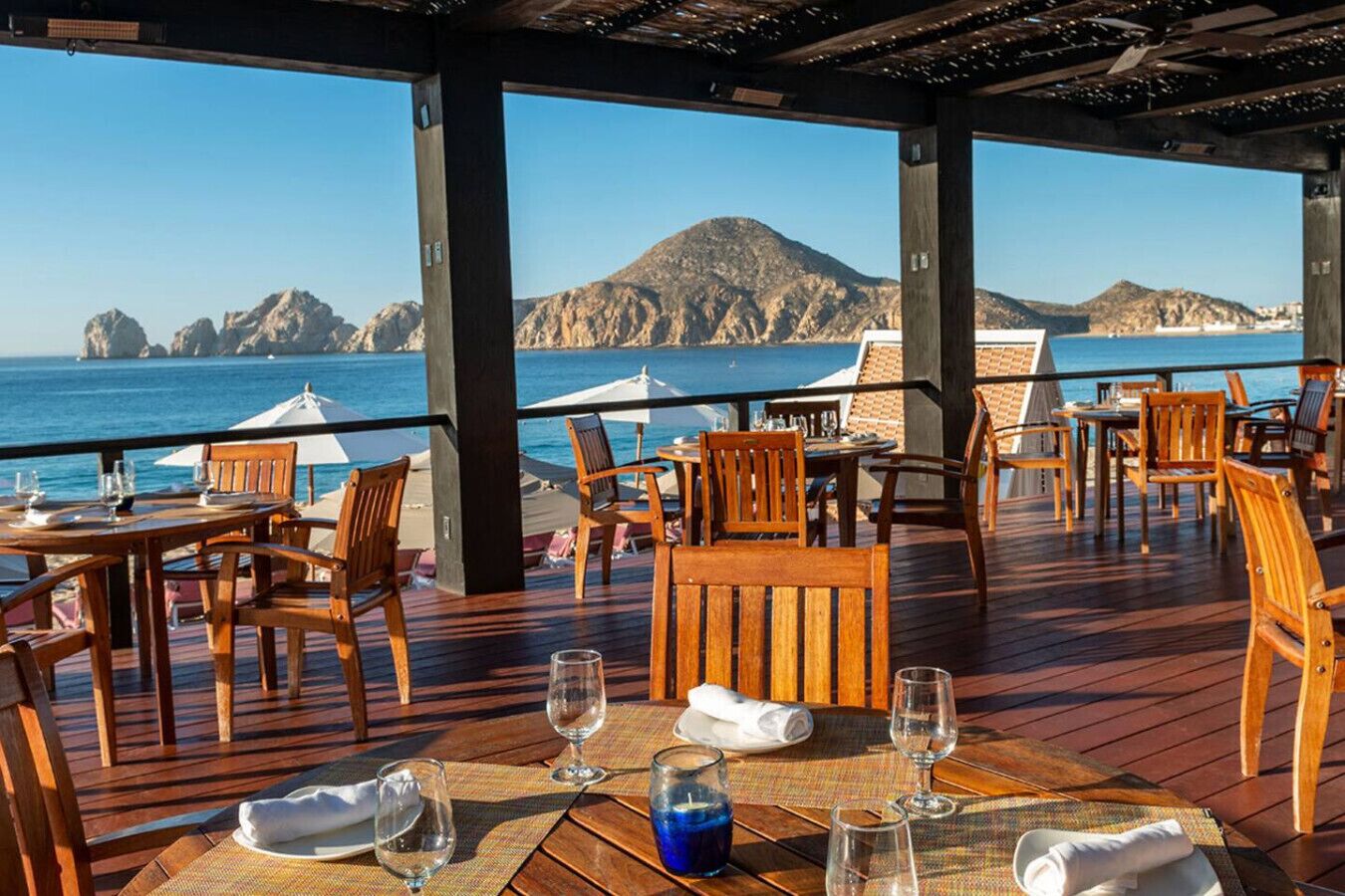 Un área de comedor al aire libre con mesas y sillas de madera, con vistas a un paisaje costero con montañas y el mar.