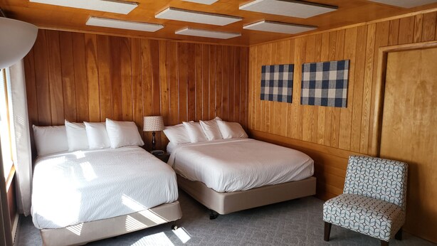 A room with two beds, a chair, wooden paneling, and ceiling lights. - Northwoods Lodge Hotel Room 2 (Long Lake)
