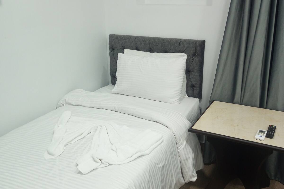 A single bed with a grey headboard, a bedside table with a remote control, and a patterned rug on the floor.
