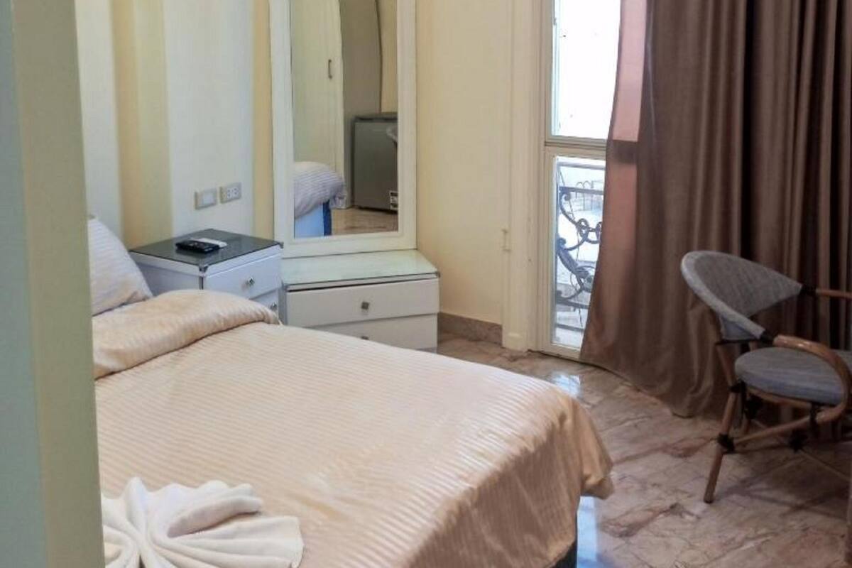 A hotel room with a bed, a bedside table with a lamp, a chair, a window with curtains, and a balcony.