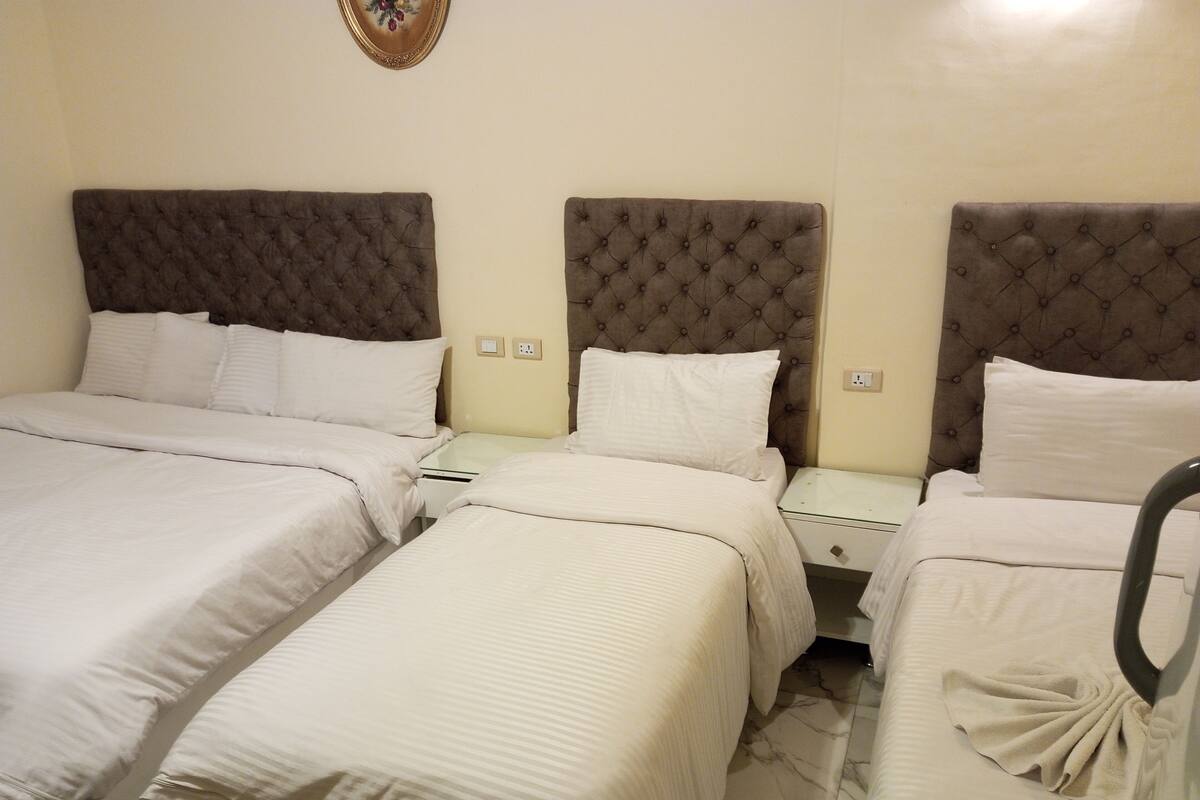 A hotel room with three beds, each with a tufted headboard and white bedding. There are bedside tables with lamps and a wall-mounted light fixture.