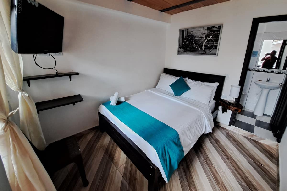Un dormitorio ordenado con una cama, un televisor montado, un estante pequeño y un baño visible a través de una puerta abierta.