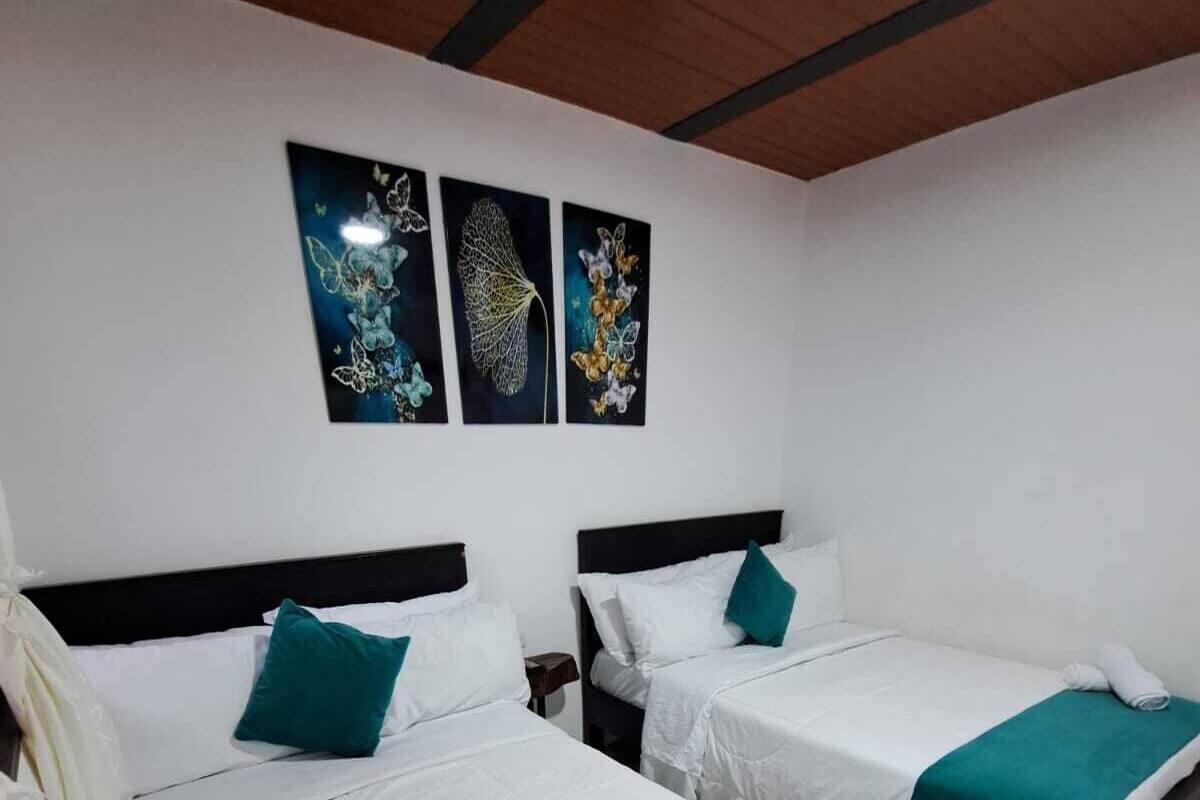 Habitación de hotel con dos camas, ropa de cama blanca, almohadones color esmeralda y una obra de arte en la pared.