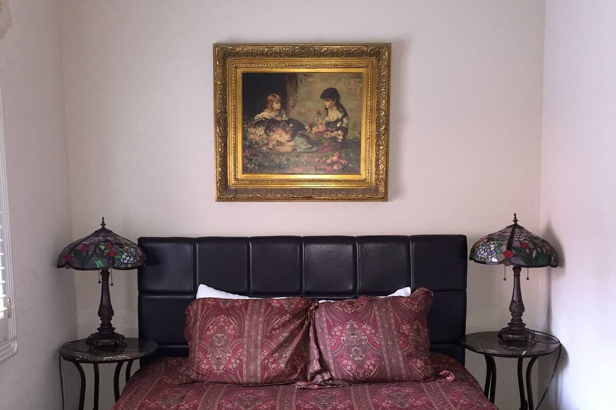 A bedroom with a bed, two nightstands, a framed painting, and a window.