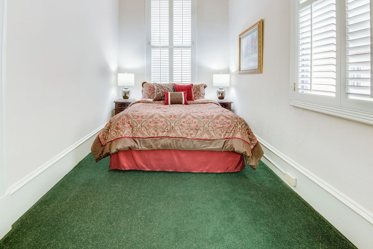 A bedroom with a large bed, two bedside tables with lamps, a framed picture on the wall, and a window with blinds.