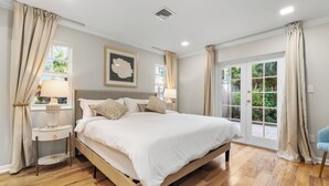 A bedroom with a large bed, two bedside tables with lamps, a window with curtains, and a view of greenery outside.