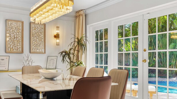 A dining area with a marble table, beige chairs, a chandelier, and a view of a swimming pool through large glass doors.