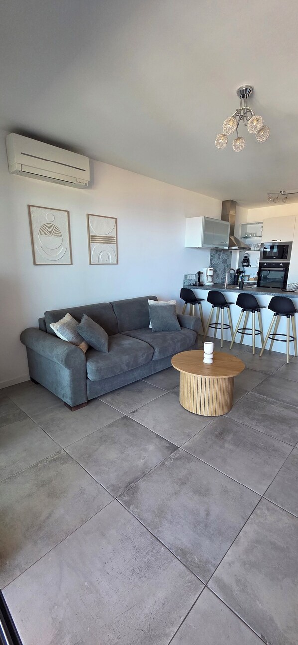 A modern living room with a grey sofa, a round coffee table, and a kitchen in the background. - Magnificent seaside apartment with terrace and sea view. (Pietrosella)