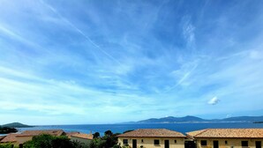 Une vue sur la côte, avec des bâtiments aux toits de tuiles rouges, un ciel dégagé et des montagnes à l’horizon.