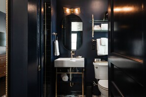 A modern bathroom with a dark theme, featuring a white sink, a mirror with lights above it, and a toilet. - The Hotel Melrose (Grand Junction)