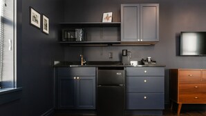 A modern kitchen with dark cabinetry, a built-in microwave, and a refrigerator. There is a wooden dresser to the right and a flat-screen TV mounted on the wall. - The Hotel Melrose (Grand Junction)