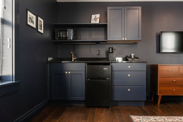 A modern kitchen with dark cabinetry, a built-in microwave, and a refrigerator. There is a wooden dresser to the right and a flat-screen TV mounted on the wall. - The Hotel Melrose (Grand Junction)