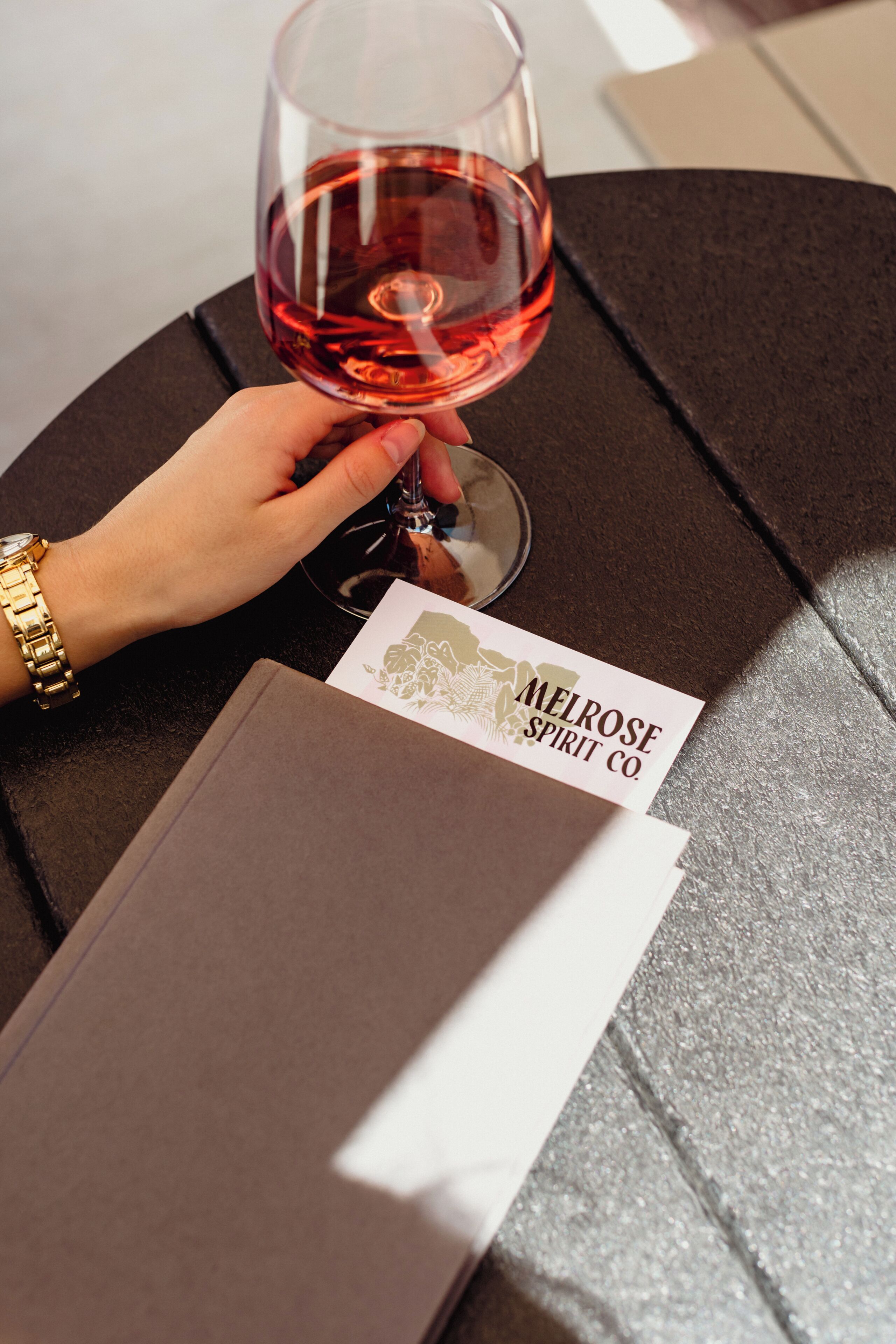 A hand holding a wine glass with a label reading 'MELROSE SPIRIT CO.' on a table with a booklet and a watch.