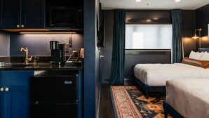 A compact hotel room with a kitchenette, a bed with a brown headboard, and a patterned rug. - The Hotel Melrose (Grand Junction)