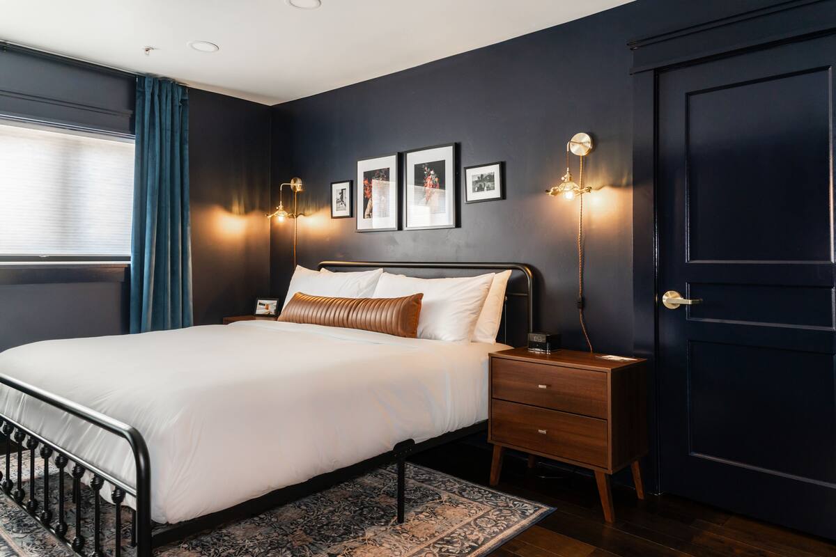 A modern bedroom with a dark wall, a large bed with white linens, a nightstand with a lamp, and framed pictures on the wall.