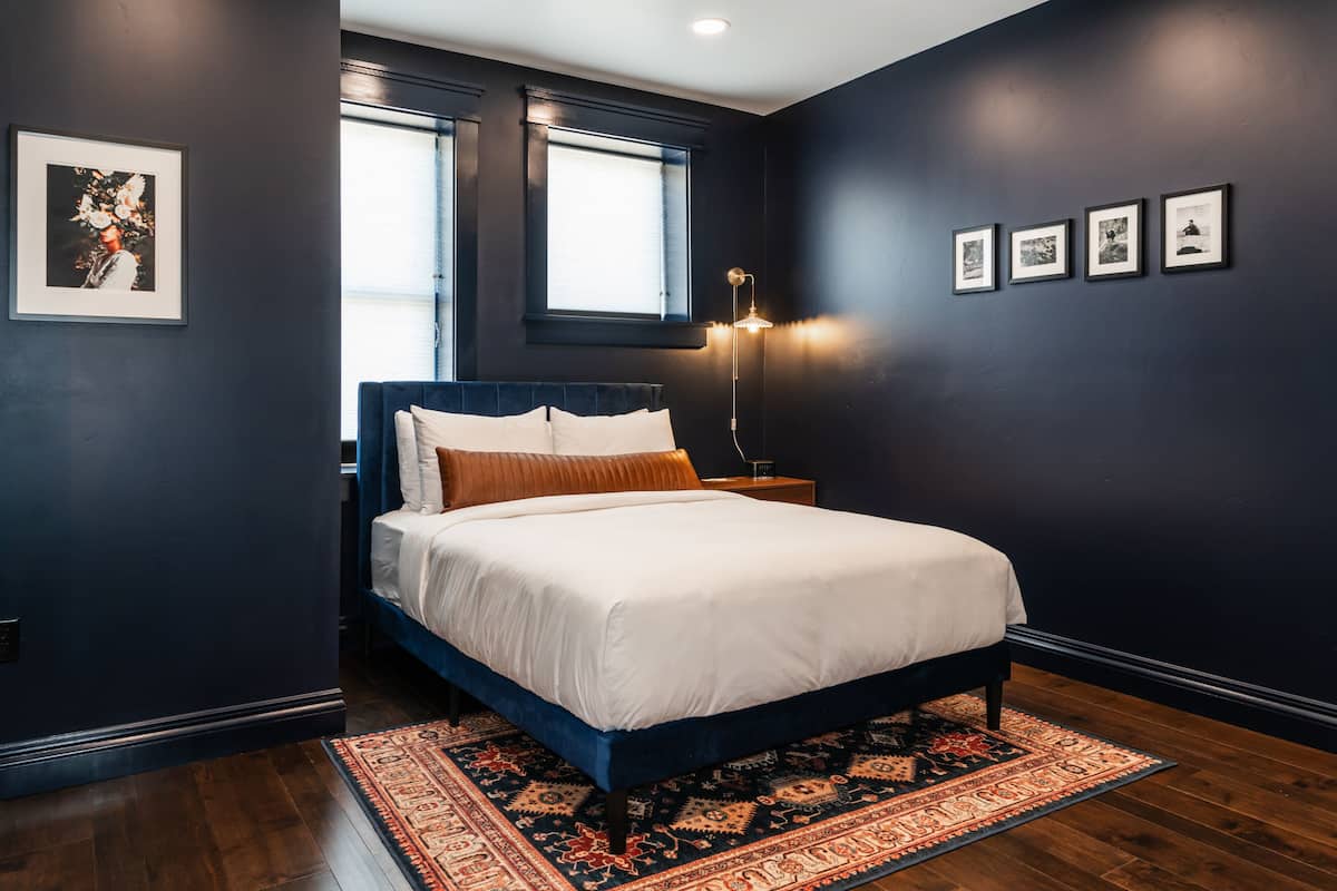 A dark-themed bedroom with a large bed, a patterned rug, and framed artwork on the walls.