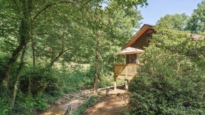 Eine rustikale Holzhütte mit einer Holzterrasse und einer kleinen Veranda, umgeben von dichtem Wald und einem Bach.