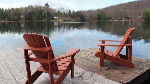Dos sillas Adirondack de madera en un muelle junto a un lago tranquilo, con árboles al fondo.