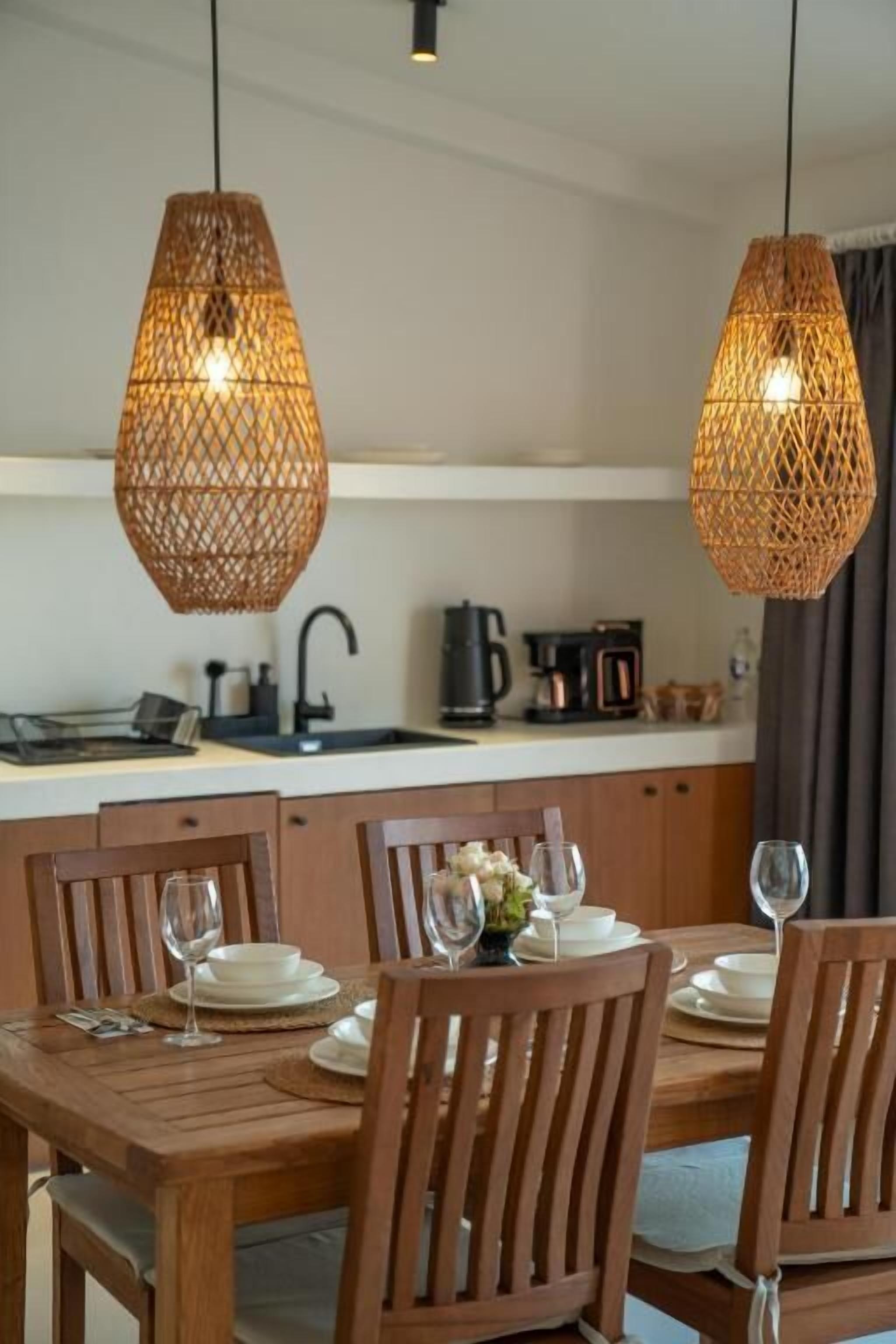 A dining area with a wooden table and chairs, two hanging wicker pendant lights, and a kitchen in the background.
