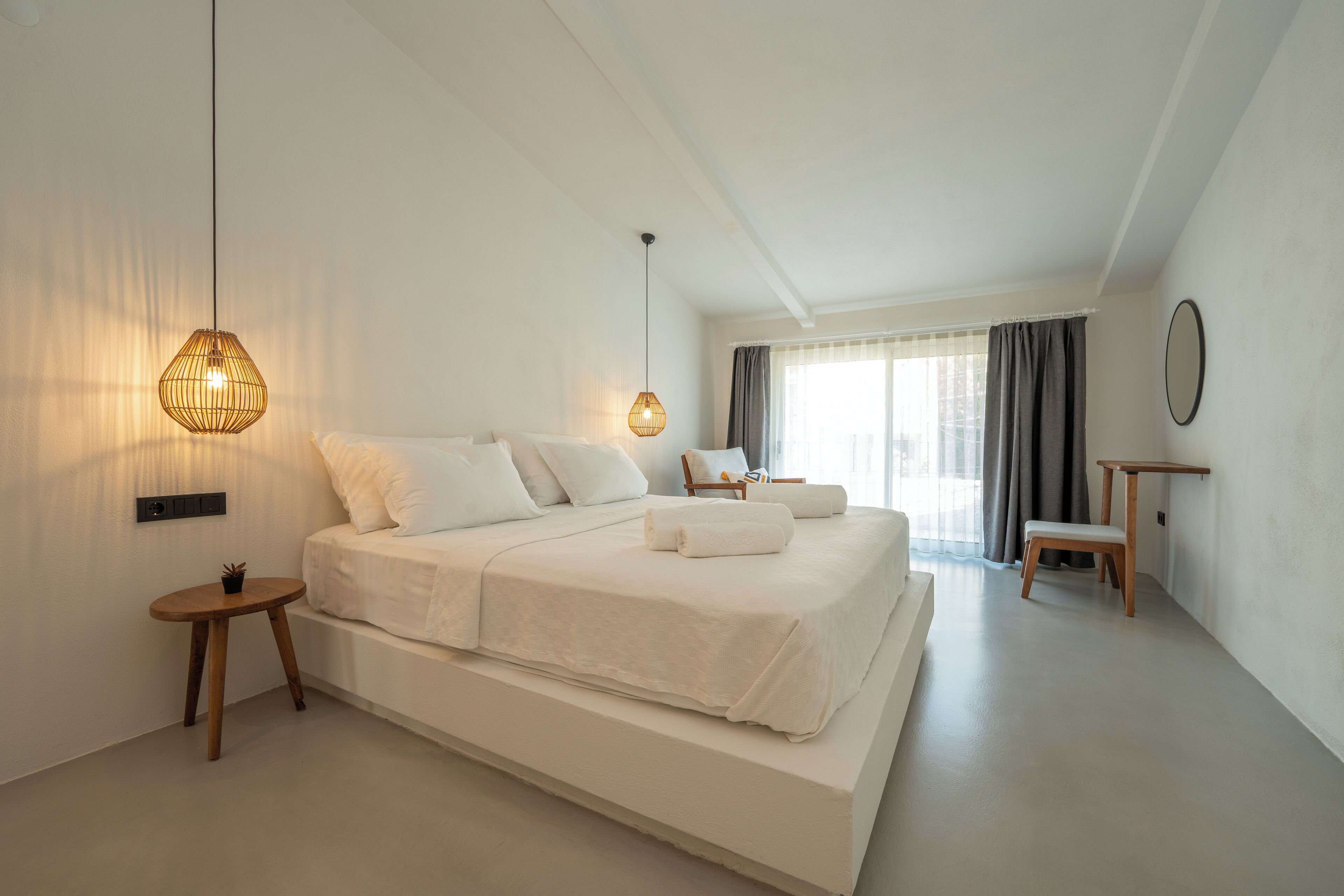 A spacious bedroom with a large bed, bedside tables, a hanging light fixture, and a view of the city through large windows.