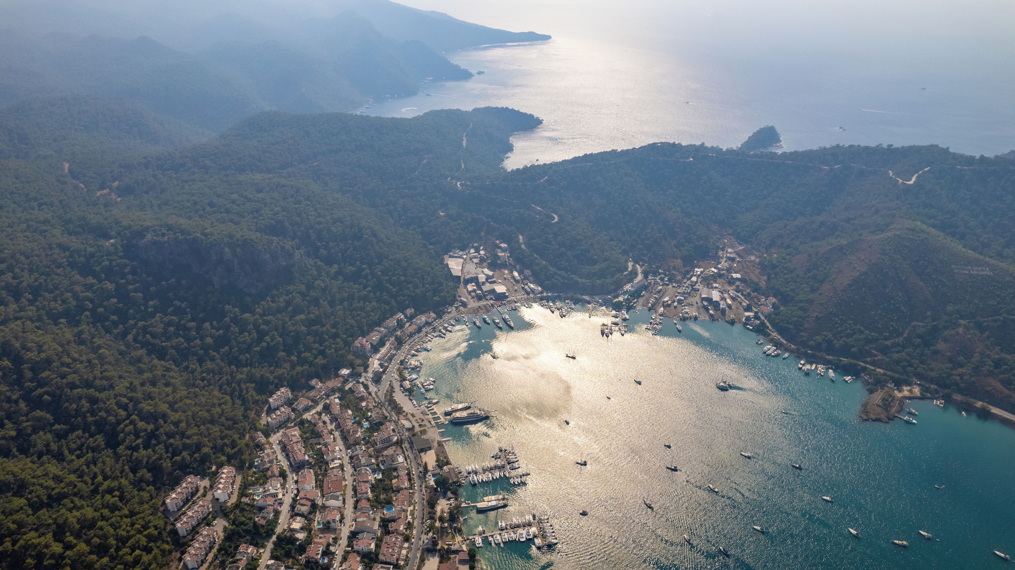 An aerial view of a coastal town with a marina, dense forested hills, and a clear view of the sea.