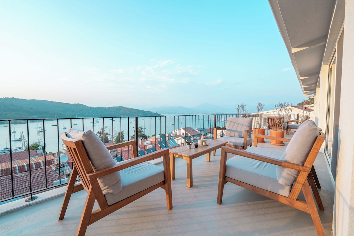 Un balcon doté de mobilier en bois, d’une table et offrant une vue sur la mer et les bâtiments.