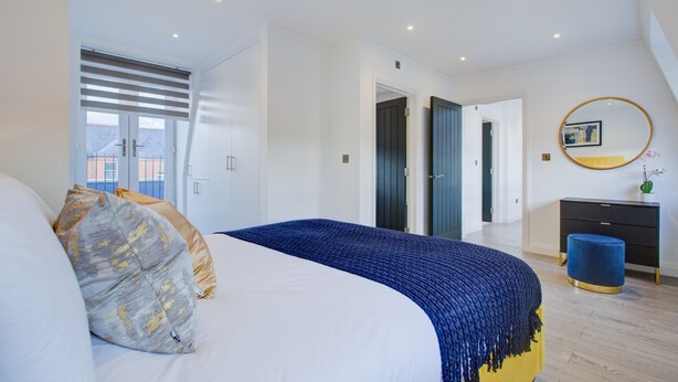 A modern bedroom with a large bed, a bedside table, a mirror, and a door leading to another room. - Hybrid Resi Fulham (London)