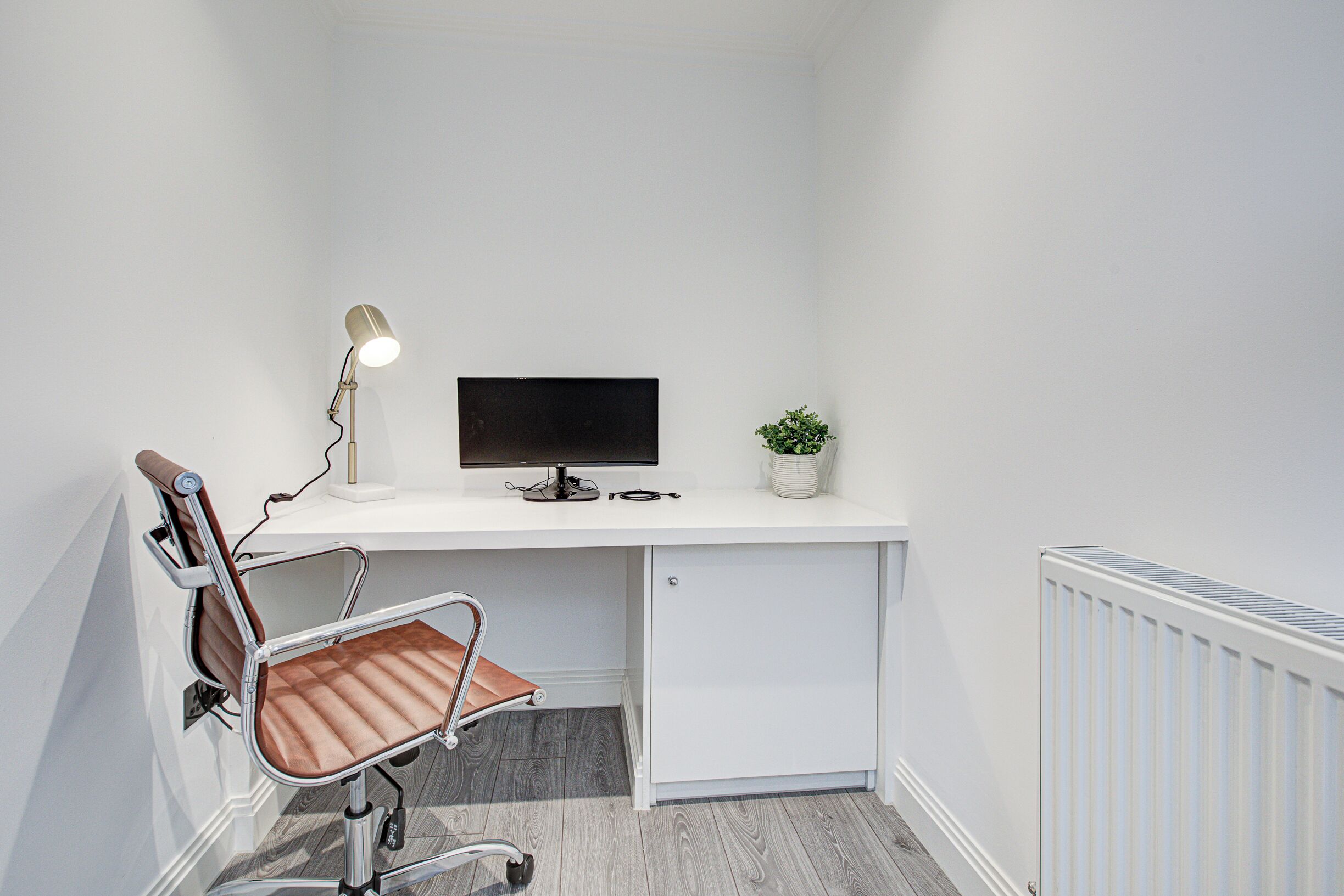 A small, white office space with a desk, chair, computer monitor, lamp, and a potted plant.