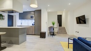 A modern kitchen with a central island, a dining area with a table and chairs, a living room with a sofa, and a staircase leading to another level. - Hybrid Resi Fulham (London)