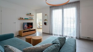 Una sala de estar moderna con televisor, sofás, un mueble bajo de madera para el televisor y un ventanal con cortinas translúcidas.