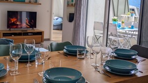 Un comedor con una mesa de madera dispuesta para dos personas, un televisor al fondo y vistas del exterior a través de un ventanal grande.