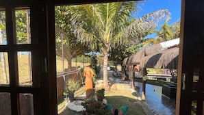 A view through a window of a tropical garden with a thatched-roof structure, a palm tree, and a statue. - Chalé Refúgio 1 (Corumba de Goias)