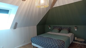 Ein Schlafzimmer mit schräg versiegelter Decke, einem Bett mit gemusterten Betttextilien, einer hängenden Leuchte und einem kleinen runden Tisch.