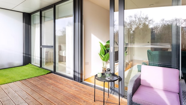 A modern room with large glass doors leading to a balcony, a wooden floor, a green mat, a pink chair, and a small table with a plant and a lantern. - Modern Chiswick Apartment (London)
