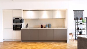 A modern kitchen with a central island, built-in oven, and a small side unit. There is a wooden floor and a plant shelf on the right. - Modern Chiswick Apartment (London)