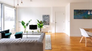 A modern living room with a sofa, coffee table, and plants. There is a television on a stand, a painting on the wall, and a dining area with a table and chairs. - Modern Chiswick Apartment (London)