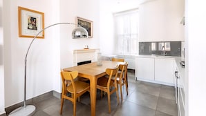 Eine moderne Küche mit weißen Schränken, einem Esstisch mit Stühlen, einer Stehlampe und einem Fenster mit Jalousien.