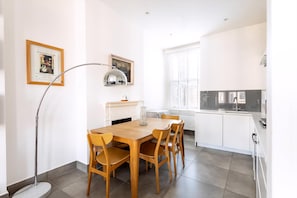 A modern kitchen with white cabinets, a dining table with chairs, a floor lamp, and a window with blinds.