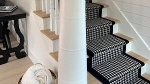 Una escalera con barandillas blancas y peldaños con estampado blanco y negro, un pilar blanco con un farol, y una cesta tejida con un objeto blanco.