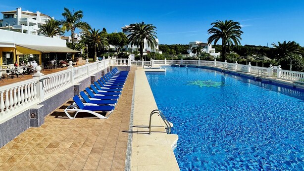 A swimming pool with blue lounge chairs and a tiled deck, surrounded by palm trees and buildings. - Elviria del Sol ground floor apartment (Marbella)