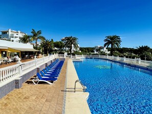 En swimmingpool med blå solstolar och ett kaklat poolområde, omgiven av palmer och byggnader.