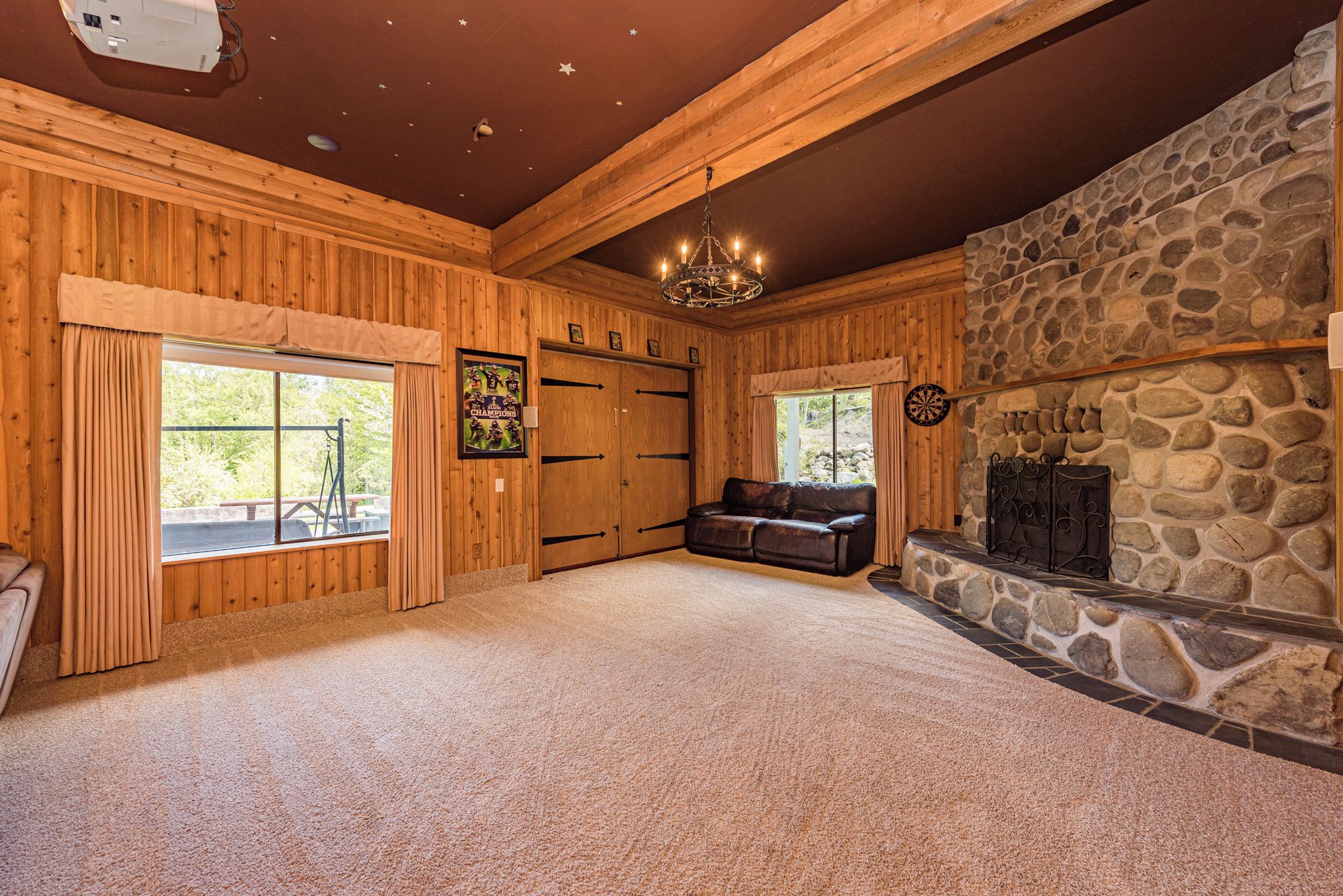A spacious room with wooden walls, a stone fireplace, a chandelier, and a large window.