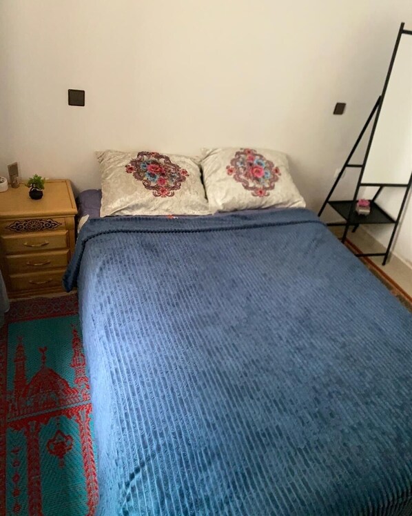 A bed with a blue blanket, two pillows with floral designs, a wooden nightstand with a small plant, and a red patterned rug. - Villa Issato (Tanger)