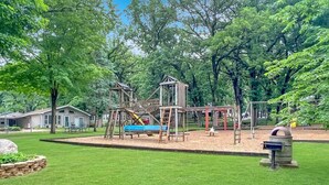 A playground with a slide, climbing structures, and a swing set in a grassy area with trees and a building in the background.