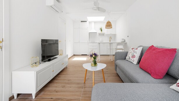Ett modernt vardagsrum med en grå soffa, ett vitt soffbord och en platt-TV placerad på ett vitt skåp.