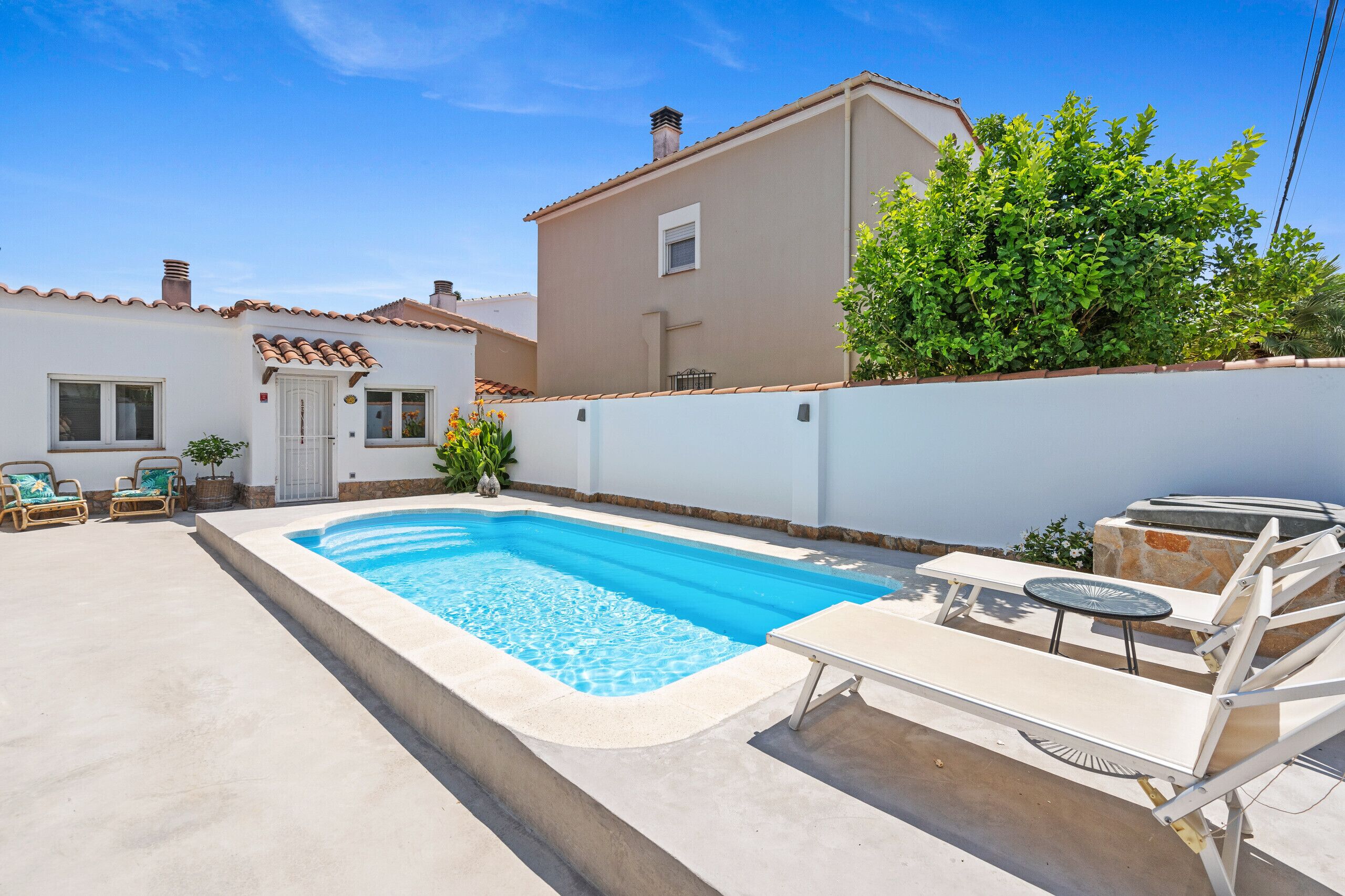 A swimming pool with lounge chairs and a table, surrounded by a white wall and a building with a tiled roof.