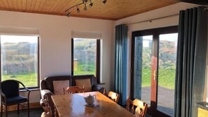 A dining area with a wooden table and chairs, a sofa, and large windows with a view of the countryside. - Luxury Sea View Cottage: Ballyconneely, Clifden, Connemara, Galway. (Clifden)