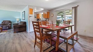 Ein modernes Wohnzimmer mit Esstisch für vier Personen, einer Couch und einer durchgehenden Tür erkennbaren Küchenzeile.