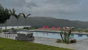 A pool area with lounge chairs and a table, surrounded by grass and palm trees, with a view of hills in the background. - Viñas del Castillo Hotel Boutique (Ensenada)